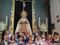 Una imagen festiva en un interior bien iluminado, con una Virgen María en el centro. Alrededor de ella, varias personas sostienen hojas blancas con texto. En el fondo, se puede ver un altar adornado con flores y velas. La atmósfera parece ser de celebración o anuncio.