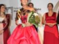 Una mujer con un vestido rojo y una corona sostiene flores, rodeada por otras personas en vestidos formales.