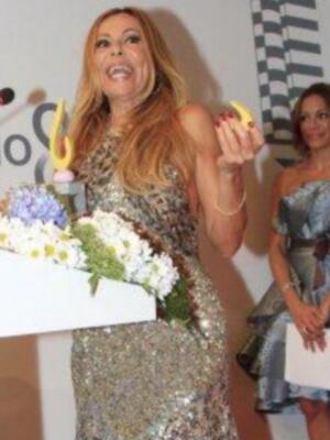Una mujer con cabello largo y rubio está en un escenario, sosteniendo una cinta amarilla con un plátano colocado en su dedo. Está vestida con un vestido brillante y tiene una corona de flores en su pecho. La otra mujer, vestida con un vestido azul, está a su lado.