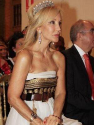 Una mujer con una corona y un vestido blanco, sentada en una silla de gala. Detrás de ella, hay personas con vestimentas formales y un hombre con una corbata roja.