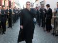 Un hombre en traje negro camina por una calle estrecha, rodeado por otros hombres también vestidos formalmente. El entorno muestra edificios con fachadas coloridas y una calle adoquinada.