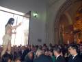 Imagen de una procesión religiosa en un templo, con una figura vestida como Jesús Cristo en el centro y una multitud de personas observando. La iglesia tiene decoraciones doradas y cristales, con una iluminación que brilla desde el fondo.