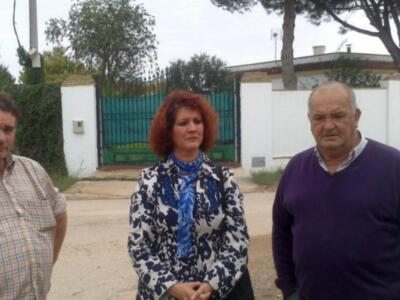 Tres personas posan frente a una casa con jardín y muro blanco.