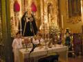 Un altar con una imagen de la Virgen María, adornado con flores y velas. En el fondo, una figura de la Virgen María en un altar dorado. Un hombre y una mujer están frente al altar, posiblemente ofrendando o rezando. La imagen está en un entorno religioso, posiblemente una iglesia.