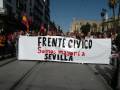 Un grupo de personas sostienen un cartel que dice "Frente Cívico Somos mayoría Sevilla" en una calle con edificios históricos y banderas.