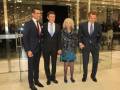 Tres hombres en trajes de negocios posan junto a una mujer vestida con abrigo y falda, frente a un edificio de cristal.