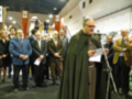 Un grupo de personas reunidas en un evento, con un hombre vestido como sacerdote al frente. La imagen muestra una reunión formal o religiosa en un lugar con carteles y luces de techo.