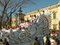 Un desfile festivo con una carroza grande y brillante, rodeada de personas vestidas en blanco. La escena se desarrolla frente a un edificio histórico con detalles arquitectónicos. El cielo es claro y azul, indicando buen tiempo para la celebración.