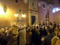 Una multitud de personas se reúne en un lugar público, posiblemente una plaza o iglesia, con iluminación navideña en el fondo. La gente está vestida con abrigos y algunos están mirando hacia arriba, posiblemente al cielo o a una estrella. La atmósfera parece festiva y el lugar está iluminado con luces navideñas.