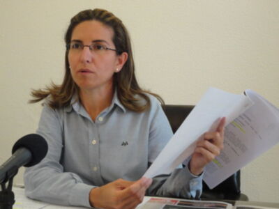 Mujer hablando en conferencia de prensa, sosteniendo documentos.