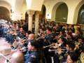 Un grupo de músicos militares toca instrumentos como trompetas, tubas y flautas en un salón con columnas y arcos. La imagen muestra una escena de concierto militar en un edificio histórico con detalles arquitectónicos.