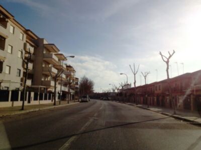 Calle desierta con edificios de dos plantas y árboles sin hojas. Un coche está estacionado en el centro de la imagen. El cielo es azul con algunas nubes dispersas.
