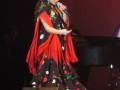 Una mujer en un vestido flamenco negro con estampado de lunares rojos y verdes, cantando en un escenario iluminado con luces rojas.
