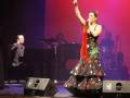 Una bailarina de flamenco en un escenario con un vestido tradicional, levantando el brazo en un gesto de triunfo, con un pianista al fondo tocando el piano.