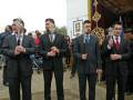 Tres hombres vestidos con trajes formales, uno de ellos sostiene un bastón ceremonial. El fondo muestra un escudo y una bandera, sugiriendo una ceremonia o evento oficial.