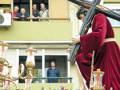 Un hombre vestido de rojo sostiene una cruz, mientras un grupo de personas observa desde una ventana. La imagen parece ser parte de una procesión religiosa, con flores y adornos dorados en el fondo.