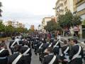 Un grupo de personas en uniforme marchando por una calle con edificios y árboles a ambos lados.