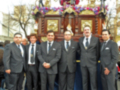 Un grupo de hombres en trajes formales se encuentra frente a una estructura histórica, posiblemente un monumento o estatua. La escena parece ser en un entorno urbano, posiblemente en una calle o plaza.