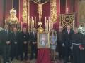 A una persona en un trono dorado con una figura vestida de blanco y rodeada por flores. En el centro, hay un crucifijo con una figura en la cruz y varias velas encendidas. A su lado, hay un cartel que dice "SANTA SEMENTA". En la parte inferior de la imagen, varios individuos vestidos con trajes formales están posando para una foto frente a esta escena religiosa.