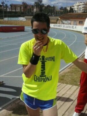 Una persona en una camiseta amarilla con el nombre "Chipiona" fuma un cigarrillo mientras se prepara para una carrera. El fondo muestra una pista de atletismo con un muro y edificios en el horizonte.