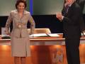 Una mujer vestida con un traje beige y una medalla colgante, junto a un hombre en traje negro, frente a una mesa con un logo de TV.
