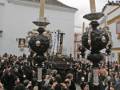 Una procesión religiosa en un entorno urbano, con personas vestidas de negro y blanco, seguidas por una figura monumental en el centro. La imagen muestra un cielo nublado y edificios blancos con detalles arquitectónicos.