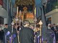 Una procesión religiosa en un entorno urbano nocturno, con personas vestidas de negro y capuchas, seguidores de una imagen venerada en un carro procesional. La escena está iluminada por faroles y la atmósfera es solemnemente festiva.