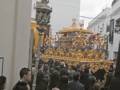 Procesión con estandarte dorado y participantes en trajes tradicionales, en una calle urbana.
