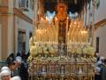 Imagen de una procesión religiosa con una imagen central iluminada por velas y adornos florales. Personas vestidas de negro forman una procesión alrededor del altar.
