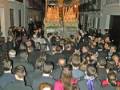 Procesión nocturna con una imagen procesional en un entorno urbano iluminado, rodeada por una multitud en trajes formales.