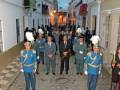 Un grupo de personas vestidas con uniformes militares caminan por una calle adoquinada en un entorno urbano. La imagen muestra una celebración o desfile militar, con algunos participantes llevando sombreros blancos y otros vestidos de manera formal. La arquitectura colonial se puede apreciar en las fachadas de los edificios a ambos lados de la calle.