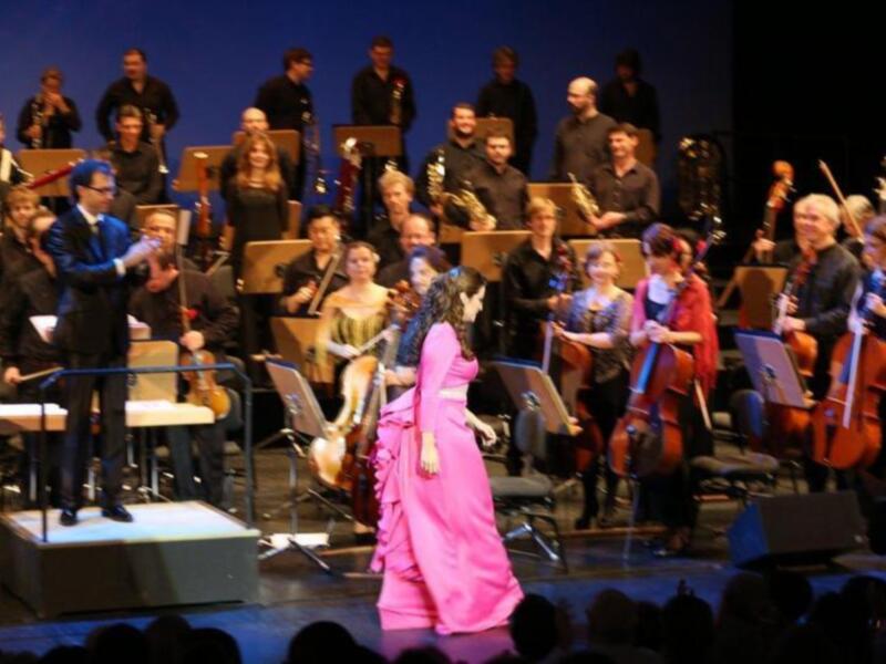 Una cantante en un vestido rosa brillante en el escenario principal, con un orquesta clásica detrás, incluyendo violines y violonchelos, en una representación de ópera o concierto.