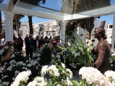 Una imagen de un cementerio con una estatua de bronce en el centro, rodeada por flores blancas y un grupo de personas observando la escena. El cielo está despejado, reflejándose en el espejo de una estructura alrededor.