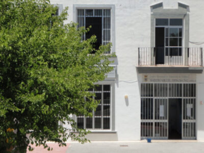 Edificio blanco con ventanas y puertas de hierro forjado, árbol grande en primer plano.