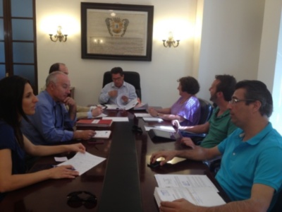 Reunión de trabajo en una sala de conferencias con miembros sentados alrededor de una mesa, sosteniendo documentos y discutiendo. La pared detrás muestra un escudo heráldico.