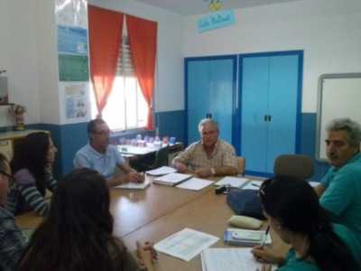 Un grupo de personas reunidas en una sala de conferencias, con libros y documentos en la mesa, y una ventana con cortinas rojas al fondo.