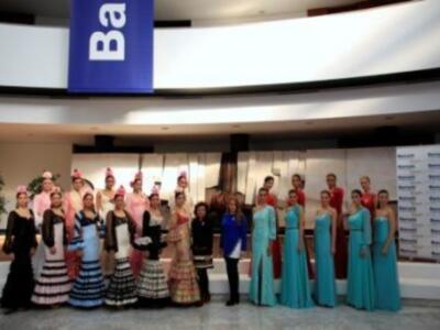 Una fila de mujeres vestidas con trajes tradicionales y vestidos largos posan frente a una pantalla gigante que muestra la palabra "Bank". La escena parece ser parte de un evento cultural o festivo.