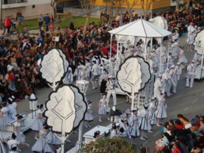 Desfile de niños vestidos con trajes blancos y sombreros azules, con un escenario central decorado con árboles blancos y negros. Público multitudinario observando el evento.