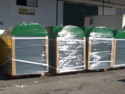 Las cajas de madera están empacadas y envueltas con papel para protegerlas durante el transporte. Se encuentran en un área de almacenamiento al aire libre, con una pared blanca y una puerta negra en el fondo.