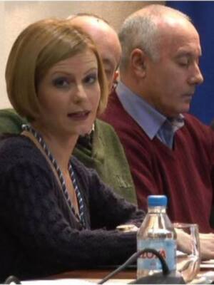 Una persona habla mientras dos personas escuchan en una sala de conferencias. La imagen muestra a tres personas sentadas en sillas rojas, con una persona en el centro hablando y las otras dos escuchando. La persona habla está usando un micrófono, mientras que las personas escuchan tienen libros y portátiles en sus manos. La sala tiene una pared blanca y un fondo con una bandera roja y amarilla.