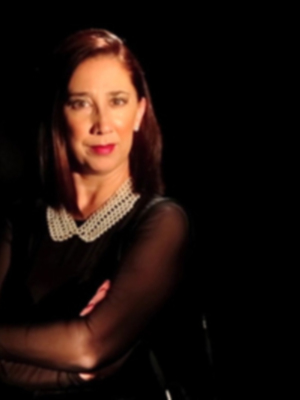 Una mujer con cabello largo y rojo, mirando hacia la derecha. Está vestida de negro con un collar de diamantes y tiene los brazos cruzados en el pecho. El fondo es negro, lo que hace que la imagen destaque más. En la parte superior izquierda hay un hashtag y en la parte inferior derecha se puede leer el nombre de una marca.