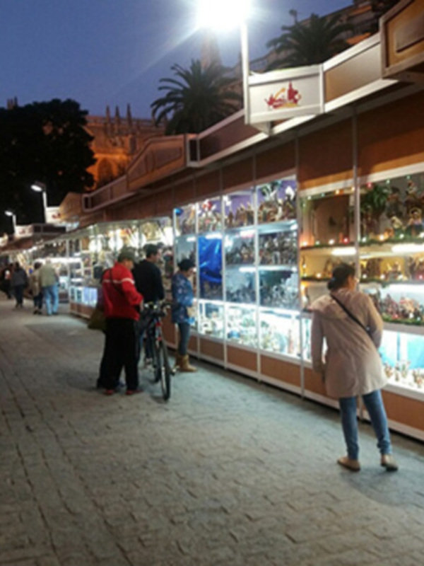 En una calle iluminada de noche, se observa a varias personas caminando y comprando en un establecimiento con vitrinas llenas de productos. La arquitectura del edificio y el cielo nocturno se pueden apreciar en el fondo.