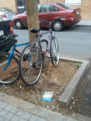 Se observa un par de bicicletas aparcadas cerca de una calle. Una de las bicicletas está apoyada en un poste y la otra está colgada del mismo. En el suelo cerca de las bicicletas hay una placa de metal con texto en inglés. Al fondo se pueden ver varios coches estacionados, incluyendo un Nissan y una camioneta roja. La escena parece ser en un área urbana con edificios de ladrillo a la vista.