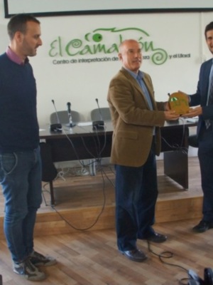Dos hombres en una sala de conferencias, uno entrega un trofeo a otro. El fondo muestra una pared con un cartel que dice 'El Camaleón Centro de interpretación de la zona y el Llobet'.