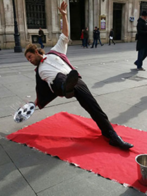 Un hombre realiza un truco de magia en la calle, lanzando una bola de cristal hacia el aire. Está sobre un tapete rojo y tiene una taza metálica en el suelo. La escena se desarrolla frente a un edificio histórico con grandes ventanas y una calle con pocos transeúntes.