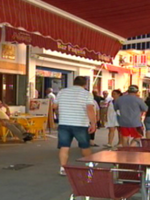 Se observa una calle con varios establecimientos comerciales, incluyendo un restaurante con toldos rojos y una tienda de ropa. La calle está llena de gente caminando o sentada en los exteriores de los negocios. La atmósfera parece ser cálida y animada, típica de un día soleado en una ciudad.