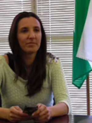 A una mujer sentada en una oficina, con un teléfono móvil en la mano. En el fondo, se puede ver una bandera nacional y persianas de ventana.