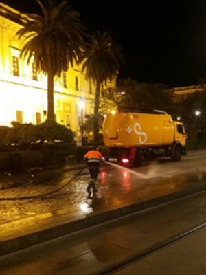 Un camión amarillo de limpieza pública rocía el asfalto con agua en la noche, iluminado por farolas y luces de edificios.
