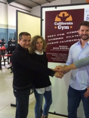 En una sala de fitness moderna, tres personas se dan la mano. La imagen muestra a un hombre y una mujer en el centro, sosteniendo las manos de otro hombre que está al lado derecho. En la pared del fondo se puede ver un cartel publicitario de "California Gym" y "Academia Paidos". La escena transmite un ambiente de colaboración o acuerdo.