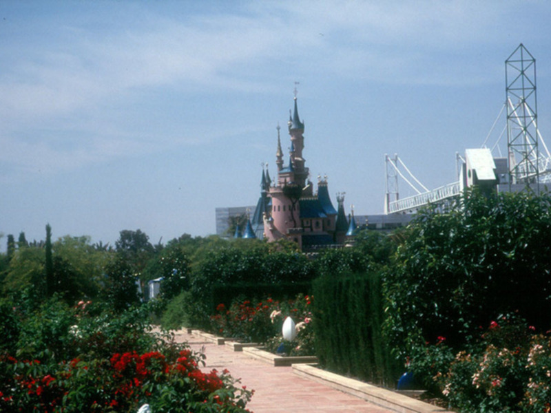 Un hermoso castillo de Disney se alza majestuosamente en el horizonte, rodeado por un parque con flores rojas y verdes. El cielo azul despejado añade un toque de serenidad a la escena.